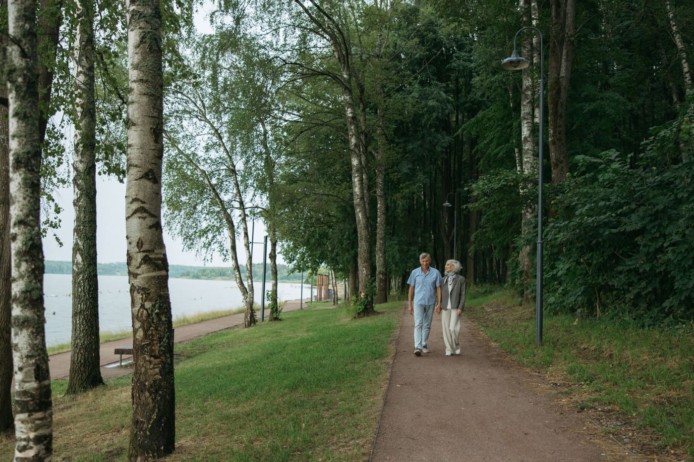 An older couple walking along a tree-lined path beside a lake