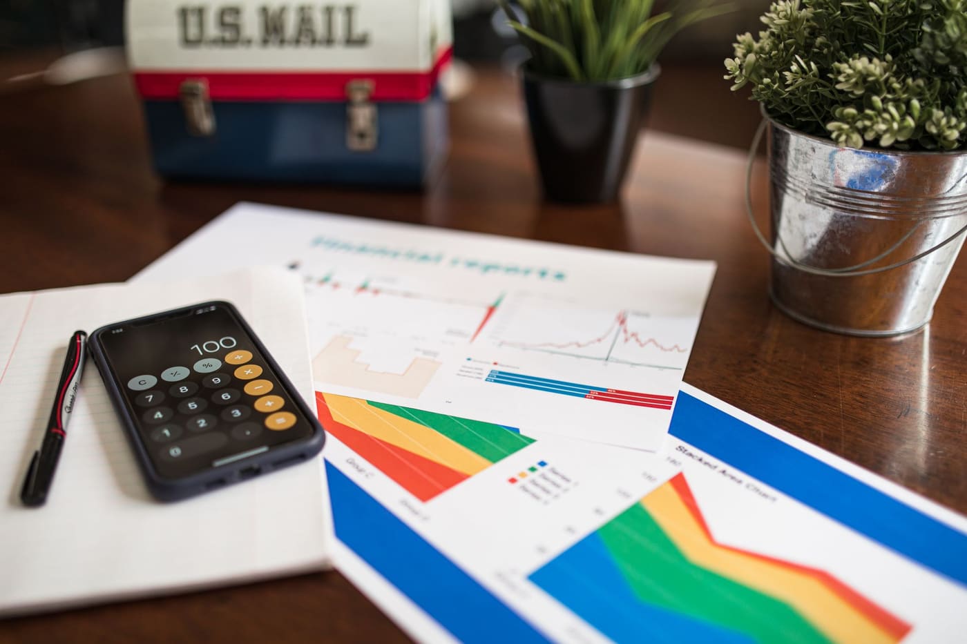 A desk with printed financial charts, a smartphone showing a calculator, a pen and a small plant