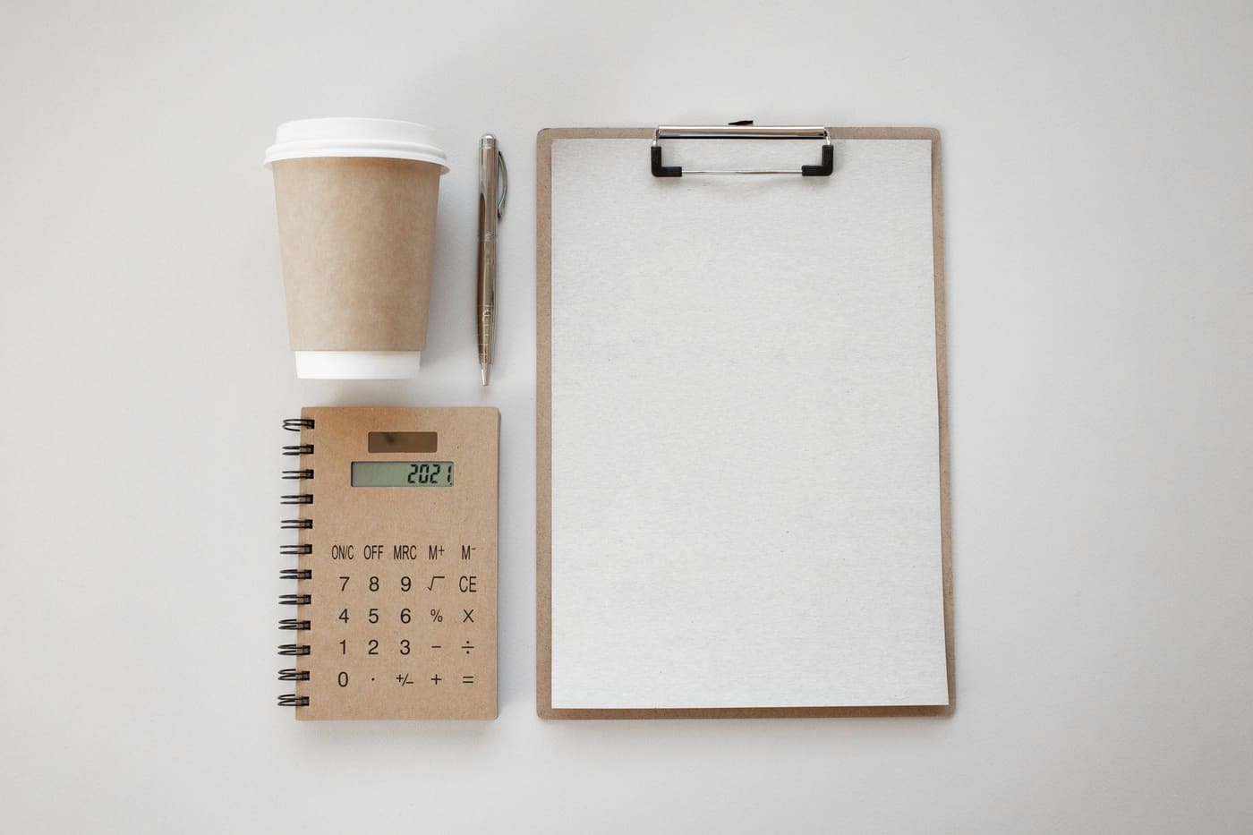 A flat lay of a spiral notebook, a small calculator, a pen and an empty clipboard on a white surface
